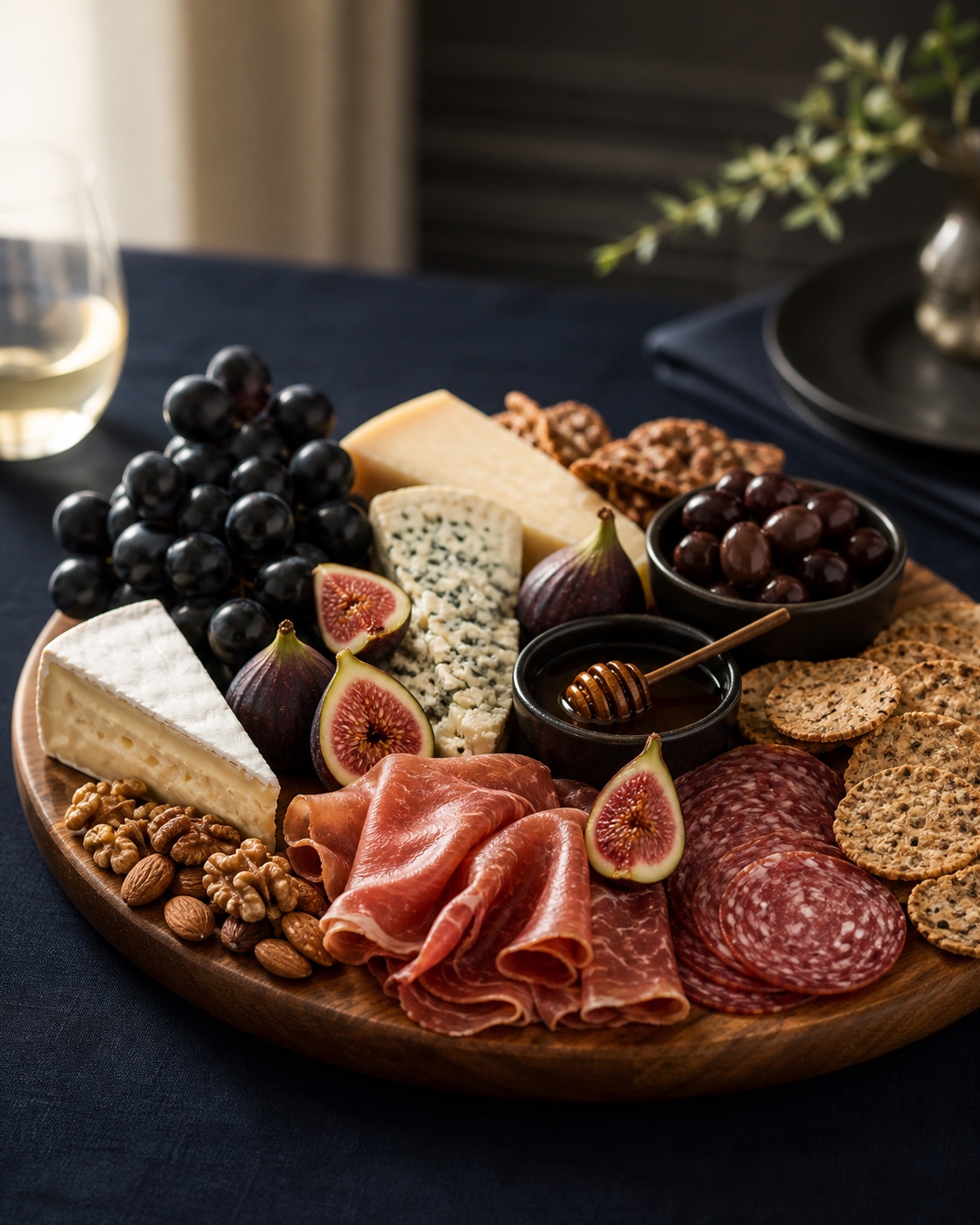 Large walnut serving board loaded with cheeses, cured meats, grapes, figs, nuts, crackers, honey, and olives on navy linen