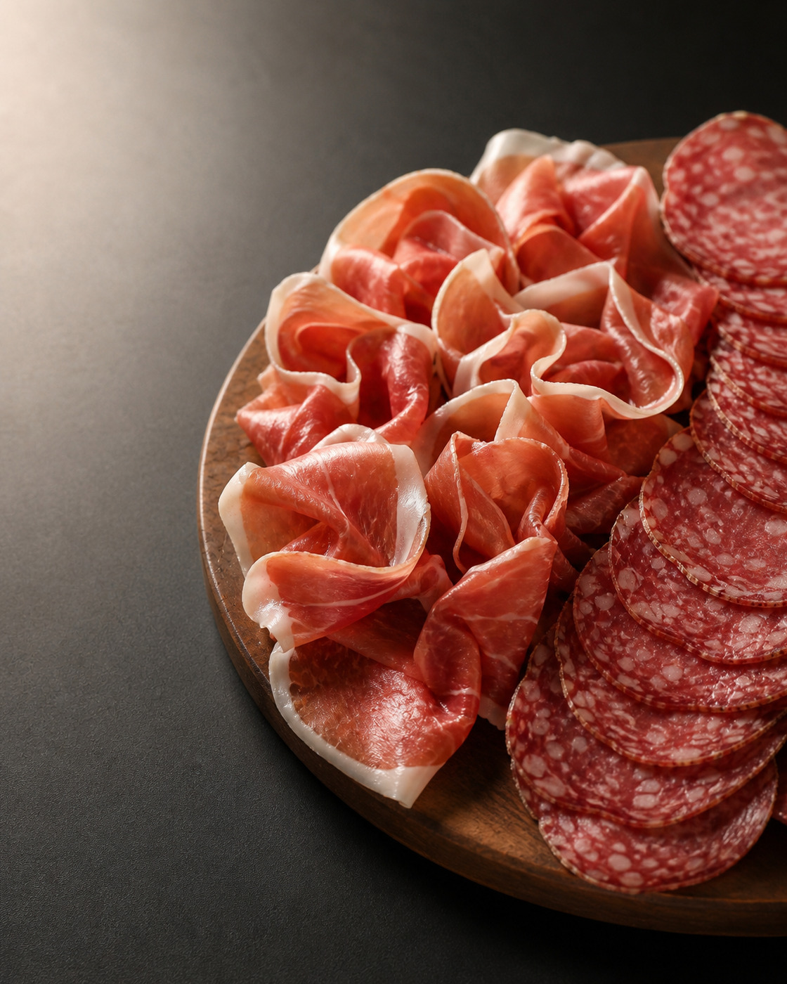 Rosettes of folded prosciutto and a fan of sliced salami on a dark surface