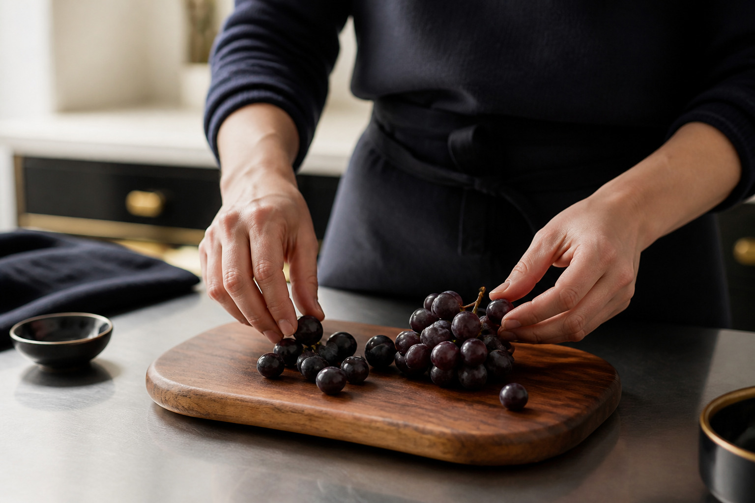 Hands arranging grapes onto a walnut board on a stainless prep counter in a commercial kitchen